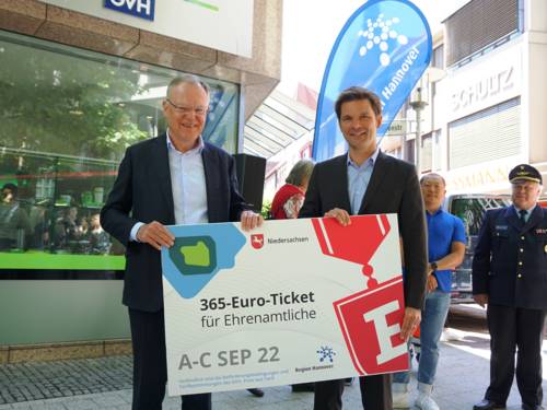 Zwei Herren halten ein großes Pappschild auf dem "365-Euro-Ticket für Ehrenamtliche" steht